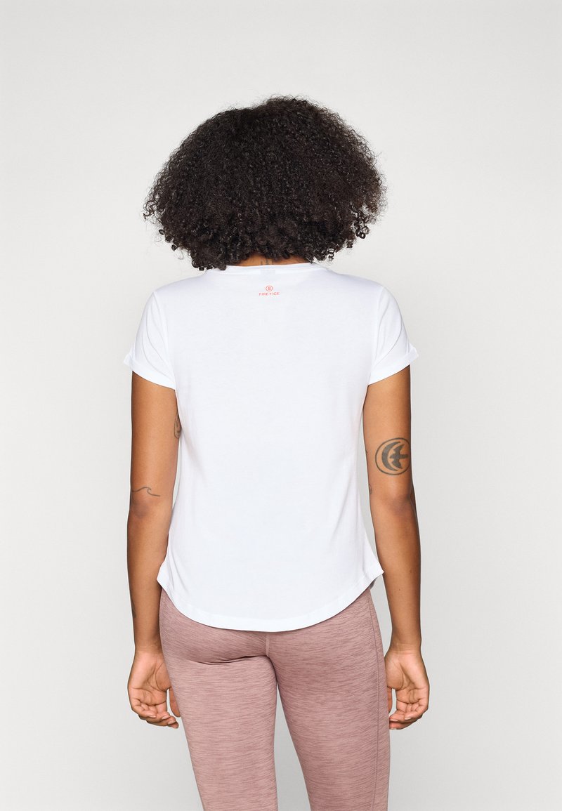 Frau mit Locken, die ein weißes kurzärmliges Hemd und helle mauvefarbene Leggings trägt, mit dem Rücken zu einer schlichten weißen Hintergrund.