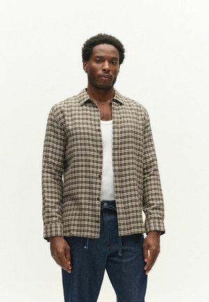 Mann trägt ein braun-weiß kariertes Hemd über einem weißen Tanktop und dunkelblauer Jeans, steht vor einfachem Hintergrund.