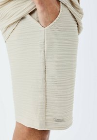 Pantalones cortos de color beige con una sutil textura acanalada, que presentan bolsillos laterales y un pequeño acento metálico. Diseño de longitud media hasta el muslo.