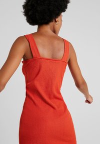 Punainen ribbitekniikalla valmistettu tankdress, jossa on leveät hihnat, neliön muotoinen selkä ja tiukka siluetti. Kangas vaikuttaa pehmeältä, luoden mukavan ilmeen.