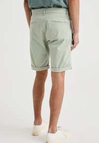 Shorts din bumbac verde deschis, cu manșete răsucite, buzunare pe spate și un accent cu logo din metal. Purtați cu adidași albi, evidențiind un stil casual.