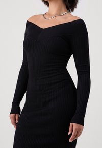 Abito lungo a maniche lunghe nero a coste con scollatura off-shoulder, silhouette aderente e tessuto testurizzato. Una collana argento aggiunge un tocco discreto.