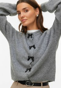 Szary dzianinowy kardigan z trzema czarnymi kokardkami z przodu, o szerokich rękawach i miękkiej fakturze. W zestawie z czarnym paskiem.