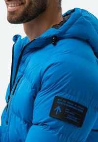 Jasně modrá zateplená bunda s kapucí, s černou nášivkou s textem na levé straně a detaily zipových kapes. Hladká textura.