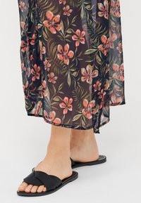 Sorte sandaler med knute-design på stroppen, kombinert med en gjennomsiktig, blomstertrykt kjole i mørke farger med rosa aksenter.
