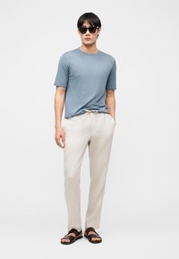 Άνδρας που φοράει μπλε κοντομάνικη μπλούζα, ανοιχτό μπεζ παντελόνι με κορδόνι, σκούρα σανδάλια και μαύρα γυαλιά ηλίου, στέκεται με το ένα χέρι στην τσέπη.