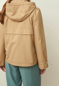 Tan waterproof jacket with a hood, featuring a bilayer design and elastic cuffs. Smooth, lightweight fabric with minimalistic detailing.