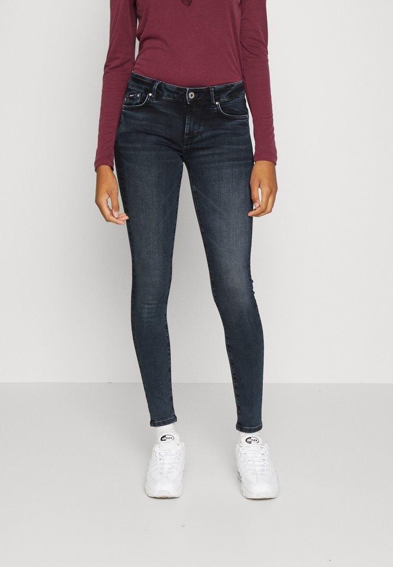 Jeans ajustados de color azul oscuro hechos de denim, con un diseño clásico de cinco bolsillos y cierre de botón en la parte delantera, combinados con una blusa color marrón y zapatillas blancas.