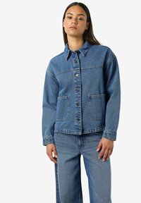 Chaqueta de mezclilla con cierre de botones, dos bolsillos frontales y un cuello clásico. Lavado medio, textura ligeramente descolorida, combinada con pantalones de mezclilla a juego.