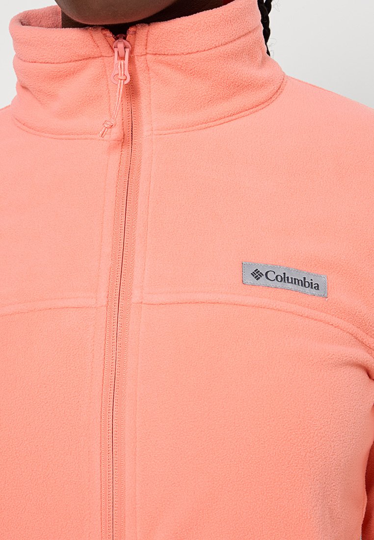 Chaqueta de forro polar rosa con cuello alto y cremallera completa. Presenta un parche del logo de Columbia en gris en el lado izquierdo. Textura suave.
