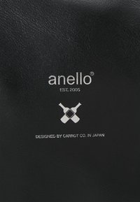 Parche de cuero negro con texto en relieve que dice "anello EST. 2005" y un gráfico de botellas cruzadas; textura suave, diseño minimalista.