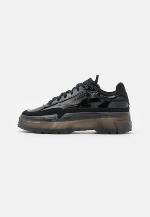 Baskets basses noires avec semelle translucide épaisse, daim texturé et panneaux en cuir verni brillant, avec détail de marque sur le côté.
