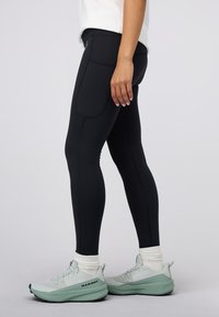 Czarne legginsy o gładkiej fakturze, z damskimi kieszeniami po bokach i wzmocnionymi szwami, zestawione z jasnoszarymi butami sportowymi.