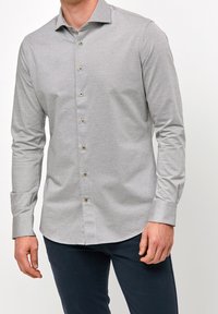 Mężczyzna w jasnoszarej koszuli w kratkę z długimi rękawami i ciemnymi spodniami, stojący na białym tle.