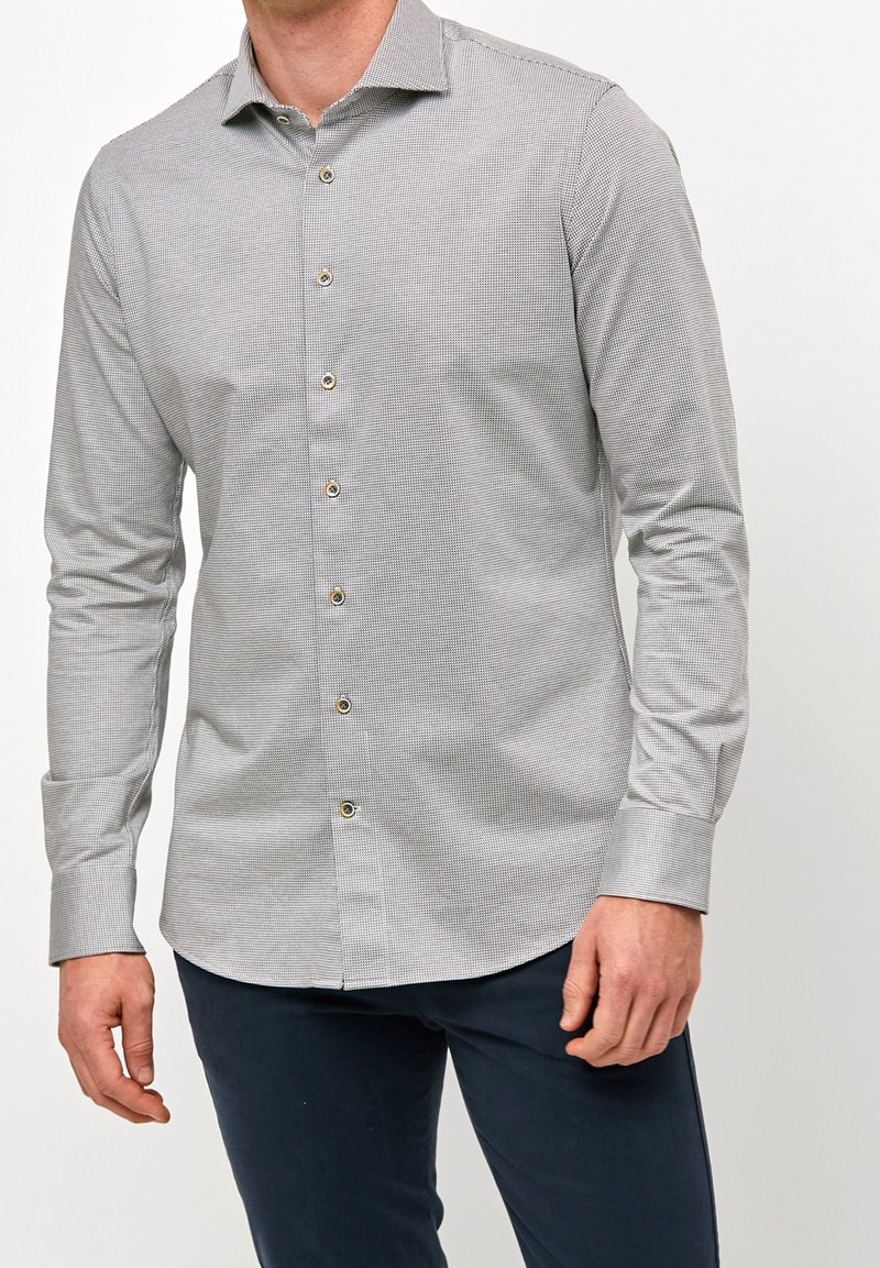 Mężczyzna w jasnoszarej koszuli w kratkę z długimi rękawami i ciemnymi spodniami, stojący na białym tle.