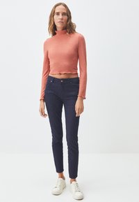 Gerippter, korallenfarbener Langarm-Crop-Top mit einem Stehkragen und gewelltem Saum, kombiniert mit dunkelblauen Skinny-Jeans und weißen Turnschuhen.