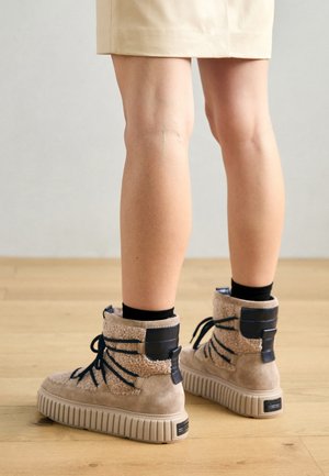 Tan suede high-top boots with textured beige accents and blue laces, featuring a thick, ridged sole and padded collar.