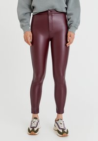 Person klädd i högmidjade, blanka burgunderleggings, grå sweatshirt och vita sneakers med lila och gröna detaljer på en vit bakgrund.