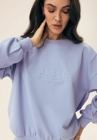 Fioletowa bluza wykonana z miękkiego materiału. Posiada ściągacze przy mankietach i u dołu, a na froncie znajduje się tłoczony napis "WEST VIRGINIA" oraz detale poniżej.