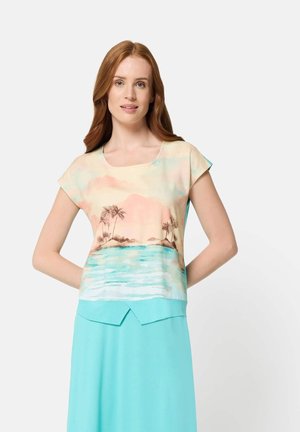 Frau mit langen roten Haaren, die ein pastellfarbenes Oberteil mit tropischem Muster und einen hellblauen Rock trägt, steht mit den Händen hinter dem Rücken.