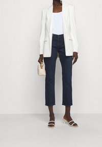 Witte blazer over een witte tanktop, gecombineerd met donkerblauwe cropped jeans en beige hoge hakken. Gecombineerd met een kleine crèmekleurige tas.