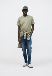 Oliwkowo zielona koszulka z logo, związana w węzeł w talii. Jasnoniebieskie jeansy, szara czapka i białe trampki z czarnymi paskami.