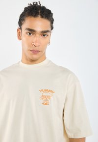 Beige short-sleeve t-shirt made from soft fabric, featuring an orange cartoon graphic with the text "YUMMY" on the left chest.