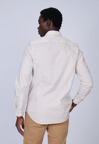 Chemise à manches longues blanche avec un petit motif géométrique rouge et bleu, dotée d'un col standard et de poignets boutonnés, rentrée dans un pantalon beige.
