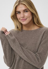 Pull marron doux avec un col rond et des manches longues, présentant un design sans coutures et un léger côtelage aux ouvertures des manches.