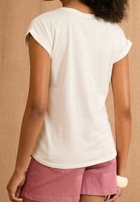 Cream short-sleeve shirt with a relaxed fit, featuring rolled sleeves. Paired with pink shorts, showcasing a smooth fabric texture.