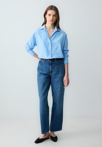 Giovane donna in piedi con una mano sulla schiena, che indossa una camicia azzurra con bottoni, jeans blu a vita alta, cintura nera e scarpe nere senza lacci.