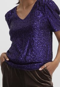 Lila paljettröja med korta puffärmar och V-ringning, med en mjuk textur och en figurnära silhuett. Matchad med bruna byxor.