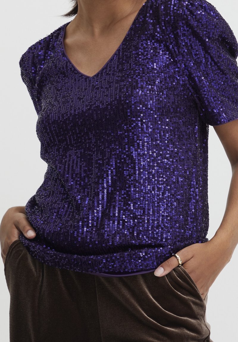 Lila paljettröja med korta puffärmar och V-ringning, med en mjuk textur och en figurnära silhuett. Matchad med bruna byxor.