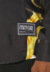 Zwarte katoenen shirt met goudkleurige en gele sierlijke accenten. Voorzien van een Versace Jeans Couture-label met decoratieve stiksels.