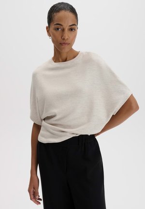 Donna con i capelli tirati indietro, in piedi con una mano sul fianco, indossa una maglia beige chiaro larga e pantaloni neri.