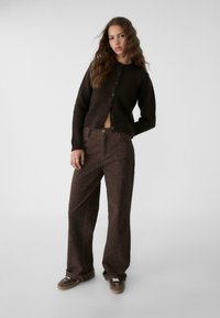 Cardigan en tricot marron avec des boutons, associé à un pantalon large marron arborant un subtil imprimé léopard. Le mannequin se tient devant un fond clair.