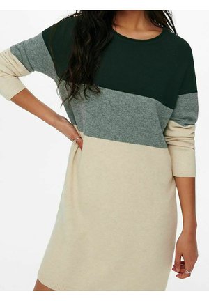 Femme portant une robe à manches longues avec des sections horizontales en blocs de couleurs vert foncé, gris et beige, la main posée sur la hanche.