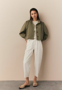 Khaki krótka kurtka z zapięciem na guziki, w połączeniu z białymi spodniami o szerokich nogawkach i beżowymi klapkami. Gładka faktura i swobodny design.