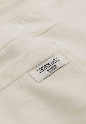 Kermanvärinen kangas, jossa on ommeltu suorakulmainen etiketti, jossa mustalla tekstillä lukee "Ainoa rajasi on mielesi" ja "Ombre vuodesta 1995".