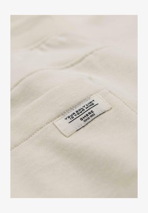 Kermanvärinen kangas, jossa on ommeltu suorakulmainen etiketti, jossa mustalla tekstillä lukee "Ainoa rajasi on mielesi" ja "Ombre vuodesta 1995".