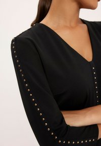 Czarny top z dekoltem w serek i rękawami o długości trzy czwarte, ozdobiony złotymi ćwiekami wzdłuż krawędzi rękawów. Gładki, lekki materiał.