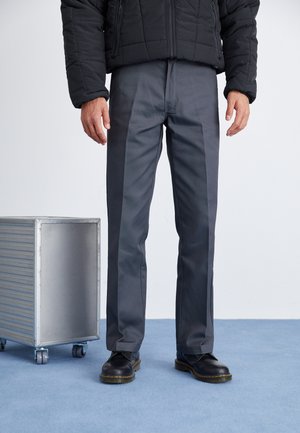 Pantalons gris sur mesure avec une coupe droite, en tissu lisse, associés à des chaussures noires, se tenant sur une surface bleu clair.