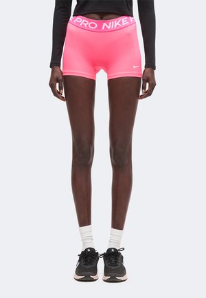 Osoba nosząca jaskrawo różowe spodenki Nike Pro, czarną bluzkę z długim rękawem, białe skarpetki oraz czarne buty do biegania Nike, stojąca prosto na jednolitym tle.