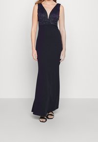 Lång svart klänning med djup V-ringning. Det övre partiet har spetsdetaljer, medan kjolen har en slät, figurnära silhuett. Bärs med svarta sandaler.