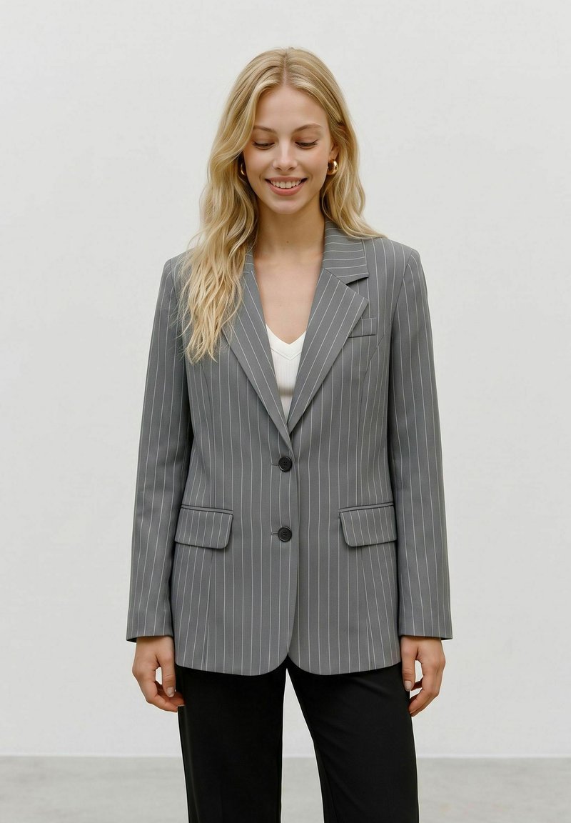 Femme blonde souriante les yeux fermés, portant un blazer gris à fines rayures, un haut blanc, un pantalon noir et des boucles d’oreilles créoles dorées sur un fond uni.