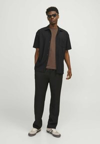 Black short-sleeve button-up shirt over a brown t-shirt, paired with loose black pants and grey sneakers with white accents. Black sunglasses worn.