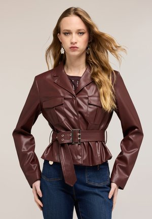 Frau mit langen blonden Haaren trägt eine weinrote Lederjacke mit Gürtel und dunkelblaue Jeans, steht vor einem einfarbigen hellen Hintergrund.