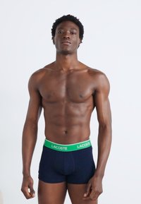 Caleçons boxers bleu marine avec une ceinture élastique verte arborant le logo "LACOSTE", fabriqués à partir d'un matériau lisse et extensible. Le modèle se tient devant un fond blanc.