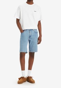 Camiseta de algodón blanca con un pequeño logo negro, combinada con pantalones cortos de mezclilla azul claros. Unos zapatos de ante marrones y calcetas blancas completan el atuendo.