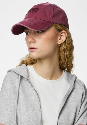 Ung person med krøllet blondt hår, iført en burgunderrød kasket, gyldne creoler, hvid T-shirt og lysegrå hættetrøje med lynlås, kigger til siden.